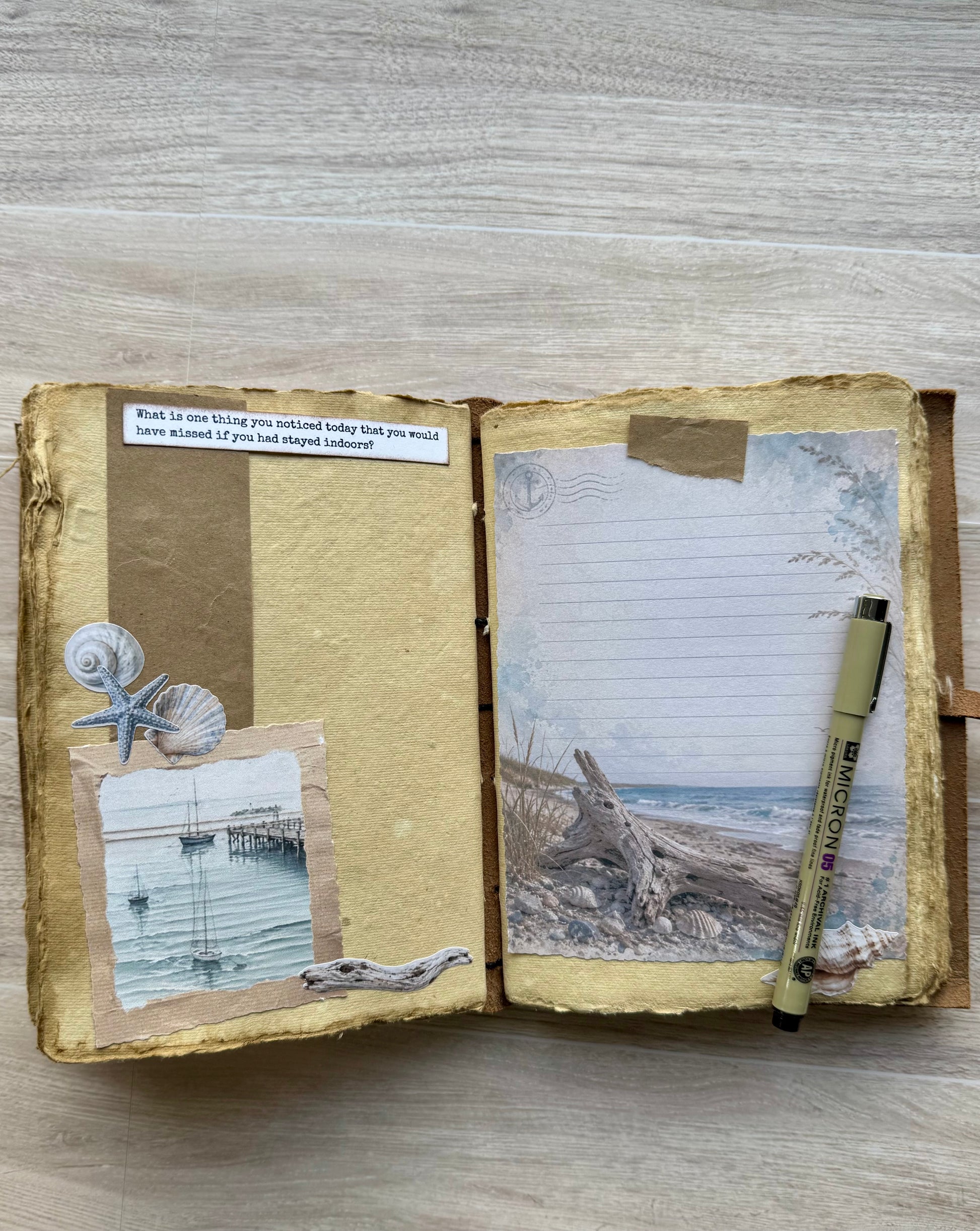 An open handmade journal with rustic, textured pages, featuring collage elements like seashells, a starfish, a small photo of a pier, a handwritten note, and a beach scene on the right page. A black and beige pen rests on the page, all set on a light wood background.