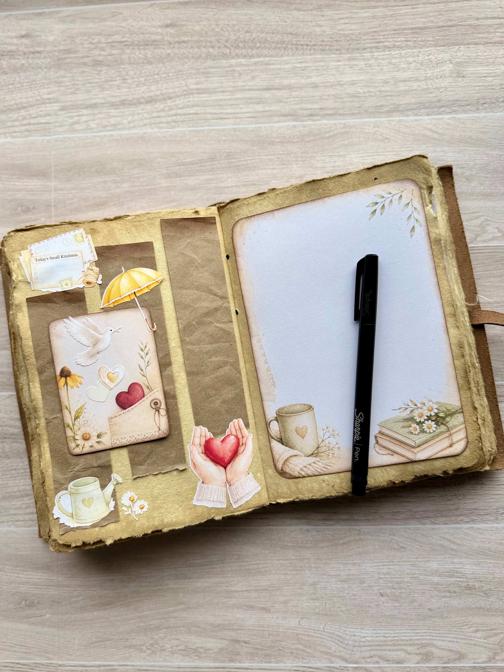 An open handmade journal with distressed brown and gold paper covers, decorated with vintage-style illustrations including a teacup, book, bird, and hearts. A black pen rests on the right page, which has blank white space, while the left page contains decorative inserts.
