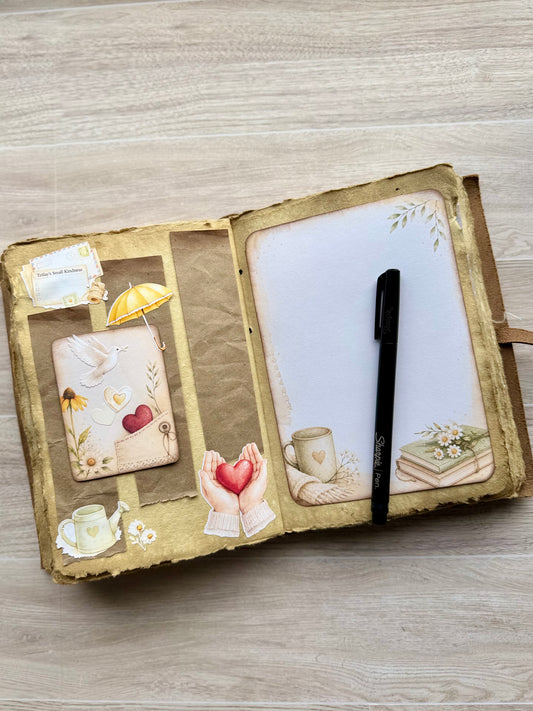 An open handmade journal with distressed brown and gold paper covers, decorated with vintage-style illustrations including a teacup, book, bird, and hearts. A black pen rests on the right page, which has blank white space, while the left page contains decorative inserts.