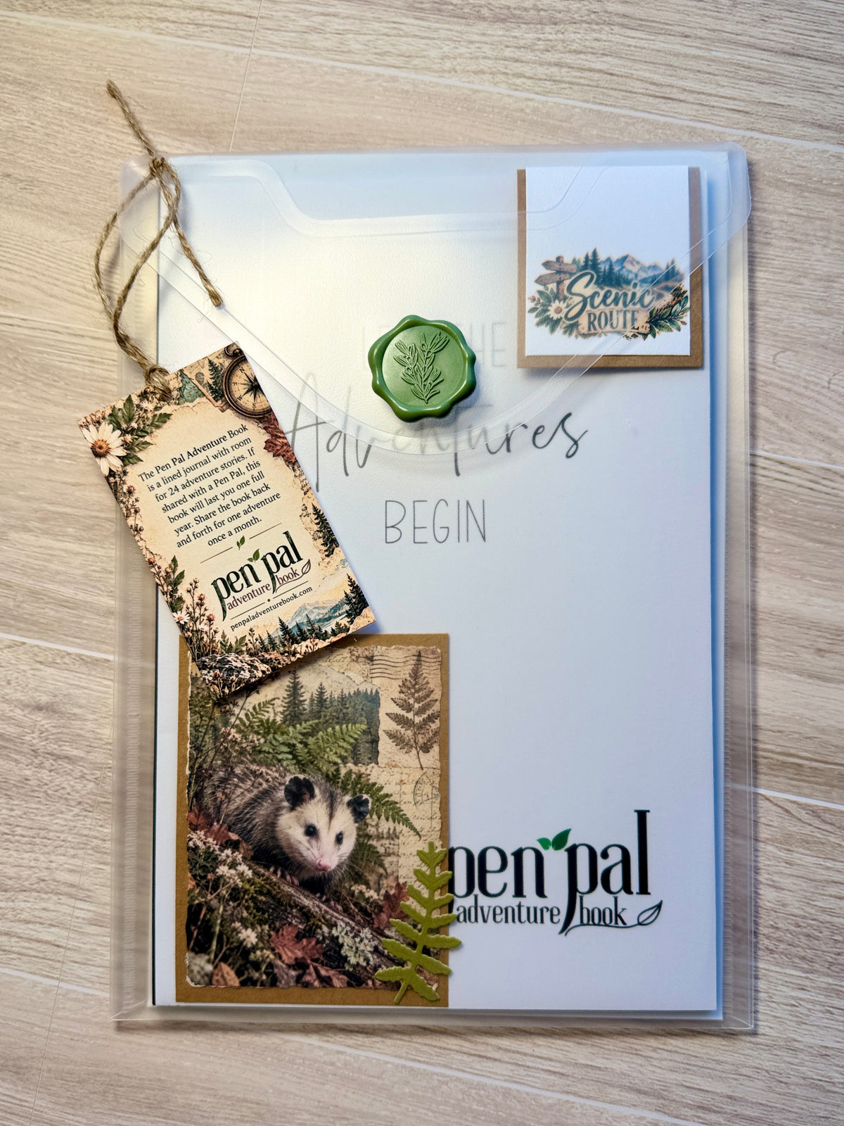 A clear acrylic book cover or folder displaying a white booklet titled 'Adventures BEGIN' with the Pen Pal Adventure Book branding. The cover includes a green wax seal, a hanging paper tag, a small scenic card, and an illustrated possum photo, all arranged on a light wood surface.
