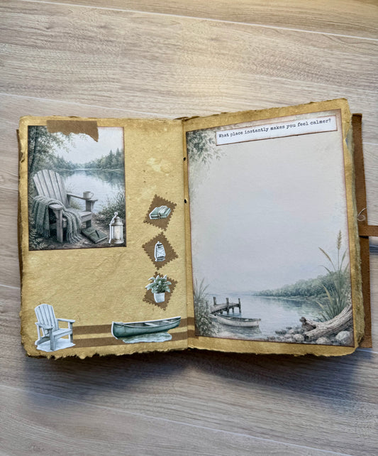 An open handmade scrapbook journal with vintage lake-themed illustrations and decorative elements, placed on a light wood surface.