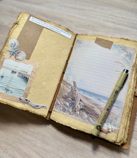 An open handmade paper journal with mixed-media collage pages, featuring a starfish, a beach scene, a piece of driftwood, a handwritten note, and a beige pen resting on the right page, set on a light wood floor.