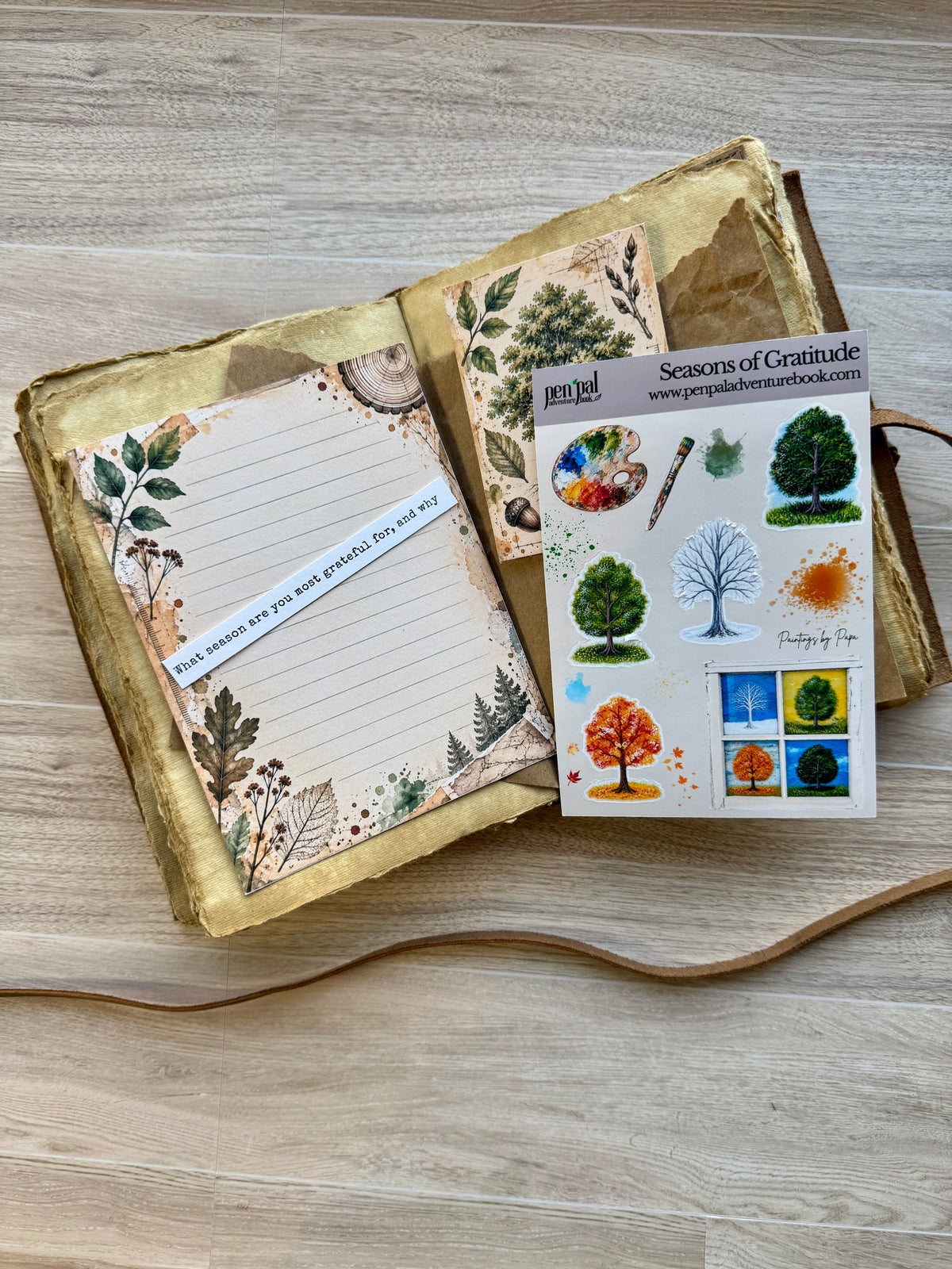 An open handmade journal with gold leaf pages and a decorative note, styled on a light wood surface next to a sheet of 'Seniors of Gratitude' paper stickers.