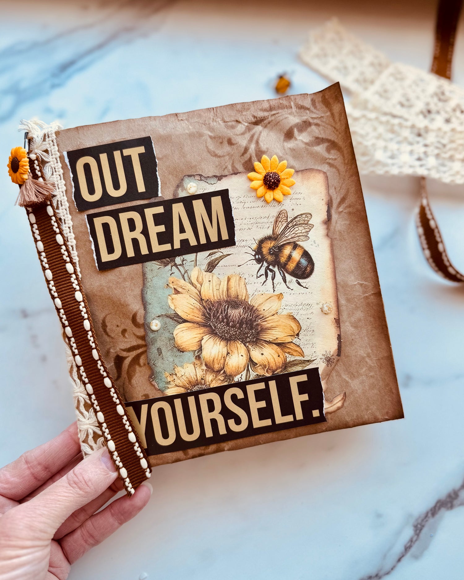 Decorative notebook with sunflower and bee design, held by a hand on a marble surface.