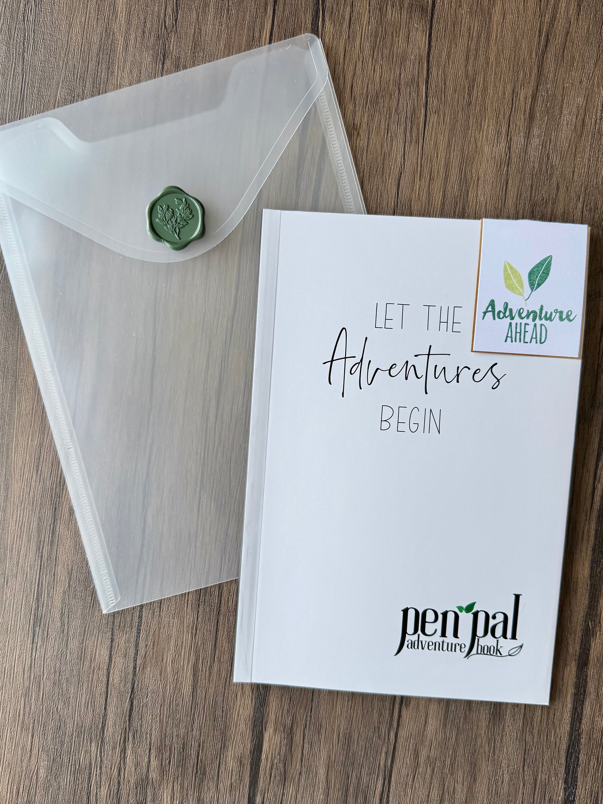 Notebook with 'Let the Adventures Begin' text and a clear envelope on a wooden surface