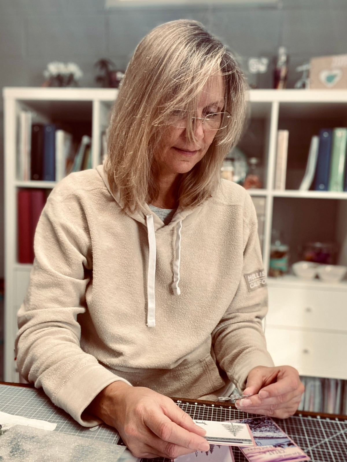 Woman sitting at a table with a neutral expression, wearing glasses and a beige hoodie.
